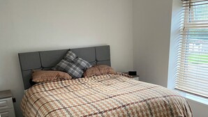 A bedroom with a grey upholstered bed, a checkered bedspread, and a window with blinds.