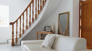 Un sofá de cuero blanco con un cojín decorativo, una escalera de madera, una mesita de madera con un jarrón y un espejo grande en la pared.
