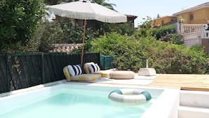 Una piscina con zona de descanso, que incluye un banco y cojines, rodeada de vegetación y una terraza de madera.