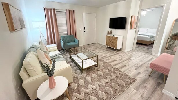 A modern living room with a sofa, a glass coffee table, a blue armchair, a TV, and a bed in the background.