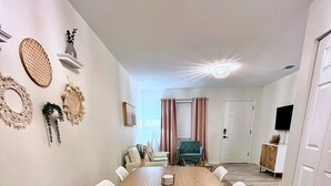A modern dining room with a wooden table and white chairs, a small side table, and decorative wall hangings.