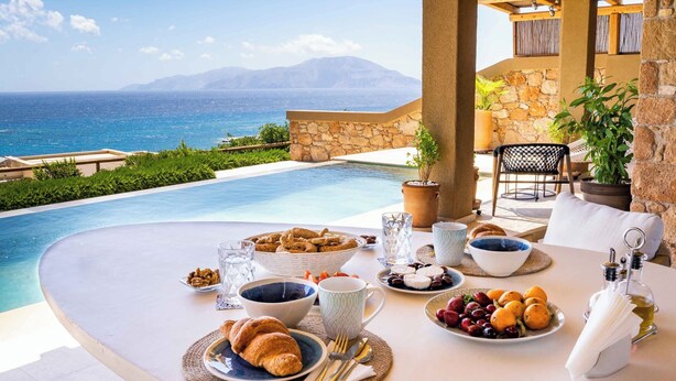 Ett frukostbord med croissanter, frukt och kaffe, med utsikt över en pool och havet.