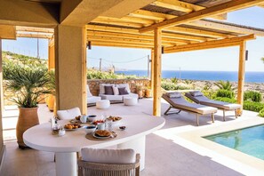 A covered outdoor seating area with a pool, stone walls, and a view of the sea.