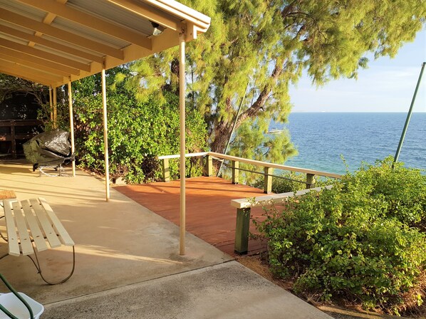 A covered outdoor area with a wooden bench, a grill, and a view of the sea.