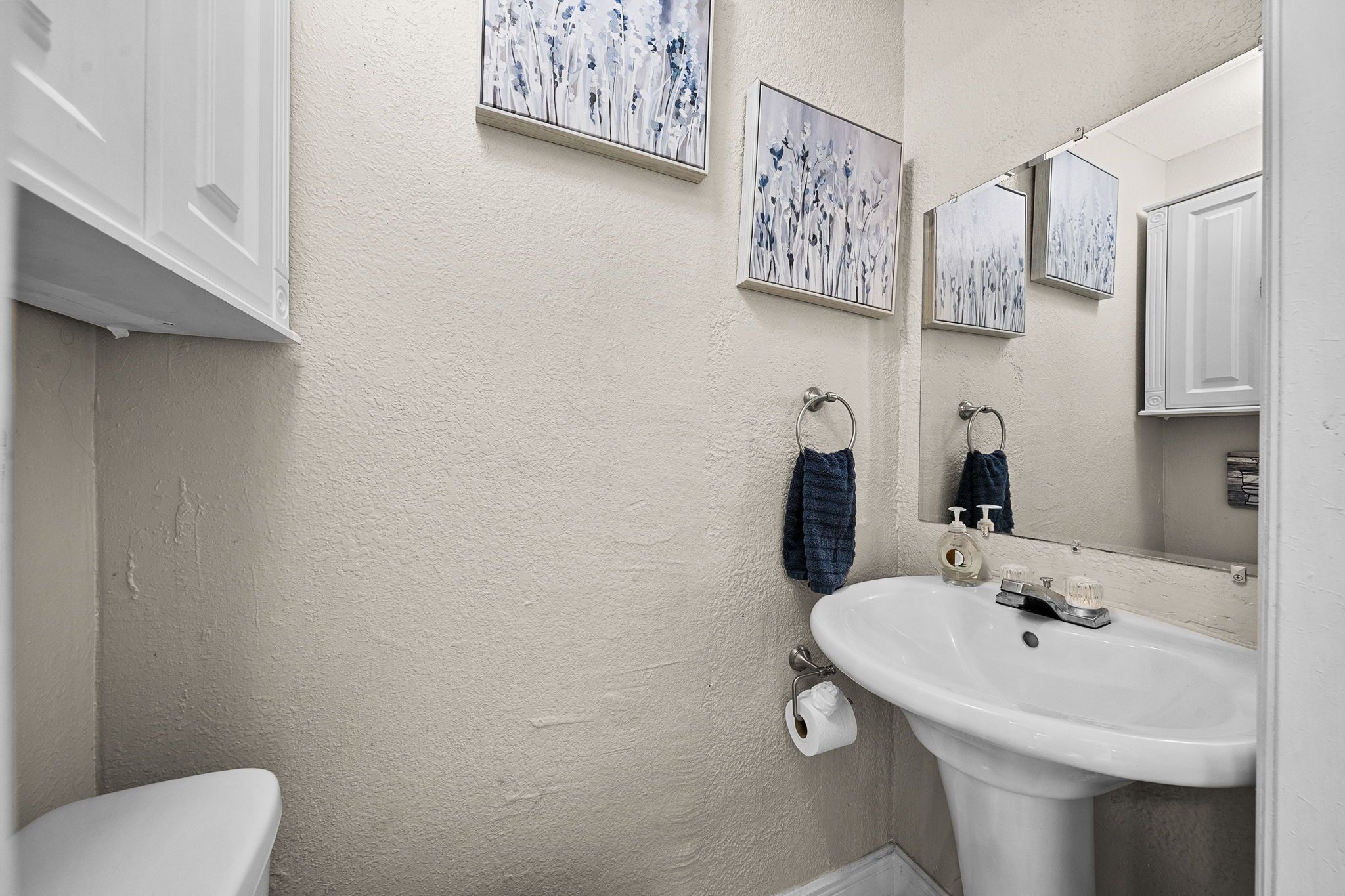 Un baño con lavamanos, inodoro y barra para toallas blancos. Hay dos cuadros con motivos florales en la pared.