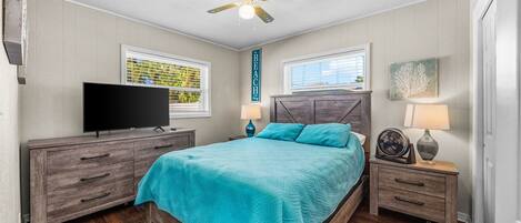 Un dormitorio con cama, ventilador de techo, televisión y dos ventanas.