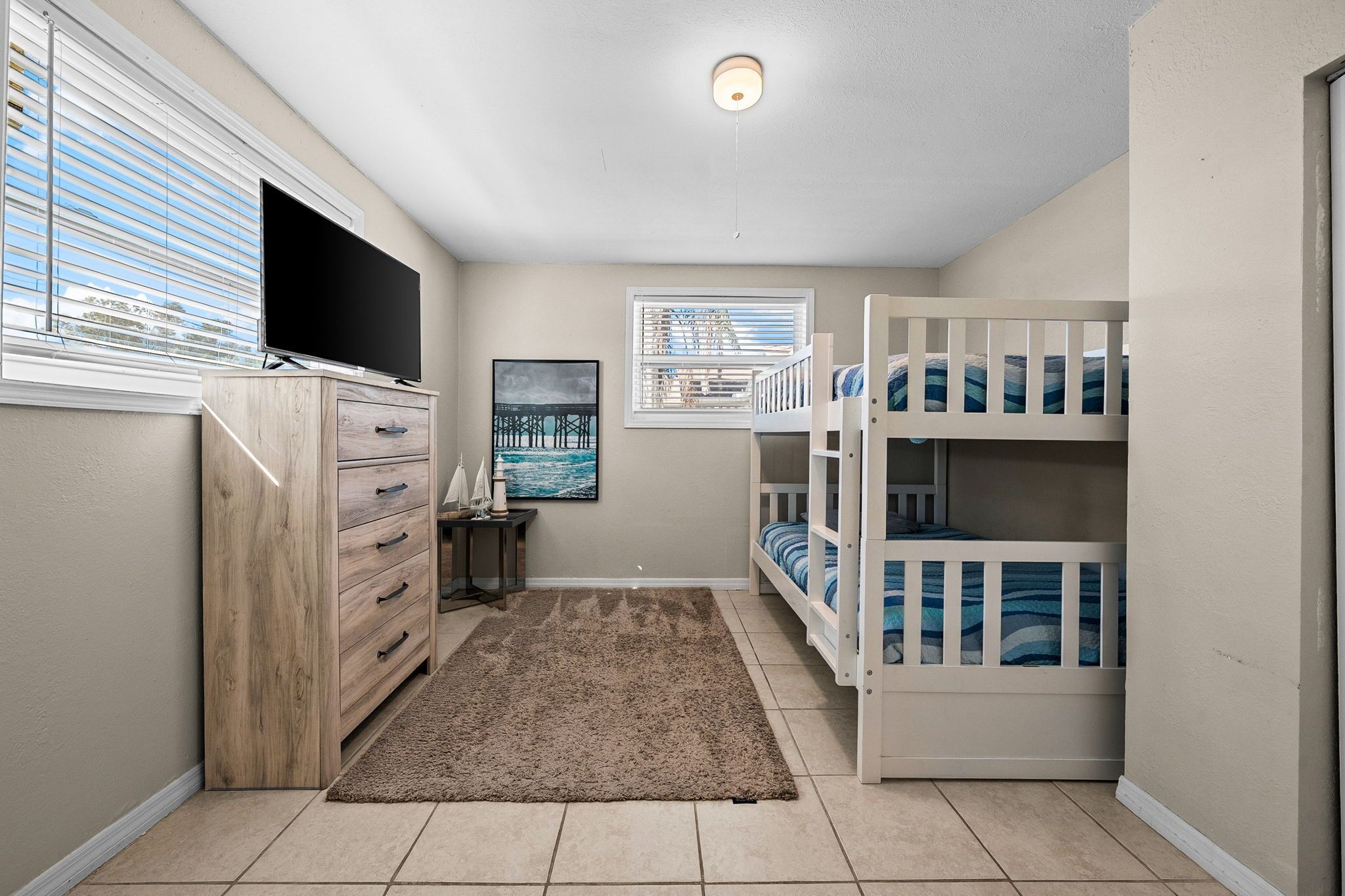 Ein Kinderzimmer mit Etagenbett, einem Holzkleiderschrank, einem Flachbildfernseher, einem Bild und einem Fenster mit Jalousien.