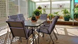 Eine überdachte Terrasse mit einem Glastisch und gestreiften Stühlen, umgeben von Topfpflanzen und einem Gitterzaun.