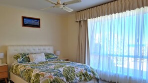Ein Schlafzimmer mit einem Bett, Deckenventilator, Nachttisch, Lampe, Fenster mit transparenten Vorhängen und einem gerahmten Bild an der Wand.