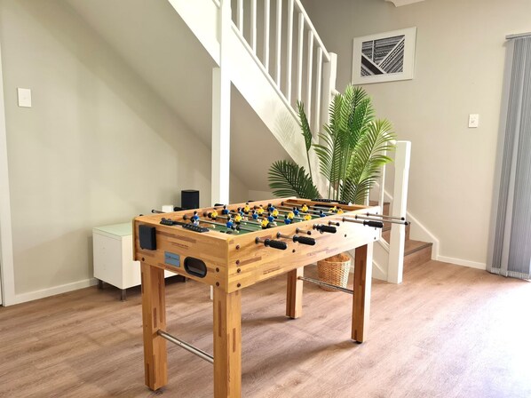 Una mesa de fútbol de madera en una habitación con una escalera, una planta en maceta y un cuadro en la pared.