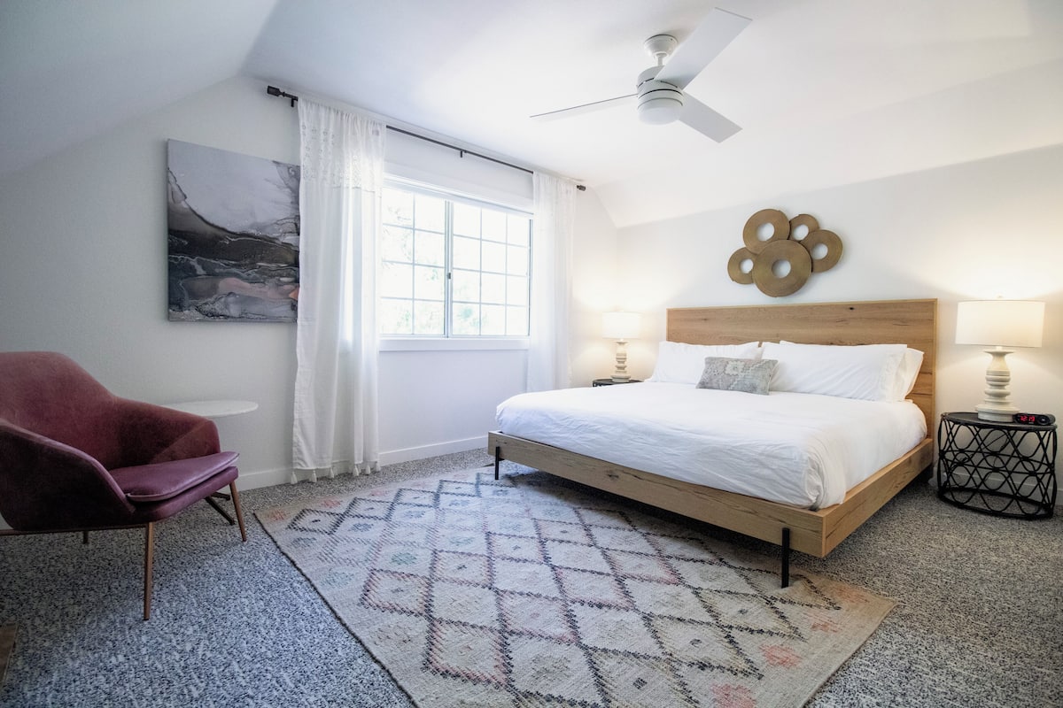 A bedroom with a bed, a chair, a window, a ceiling fan, and a wall decoration.