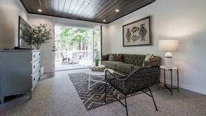 A modern living room with a sofa, a patterned rug, a side table with a lamp, and a view through an open door to an outdoor area. - Harvey House (Idyllwild)
