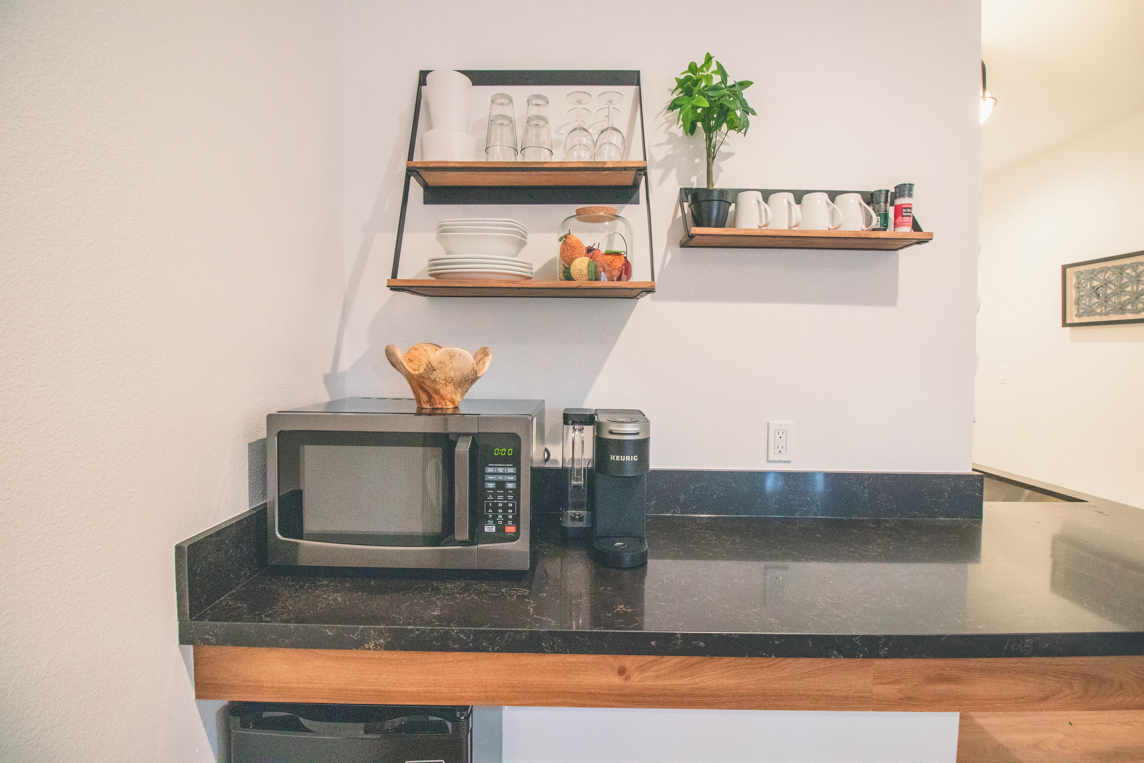 Una cocina con microondas, cafetera y estantes con vajilla y una planta.