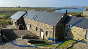 A stone building with a sloped roof, a small porch, and a view of a body of water and hills in the background. - Awel y Môr - 3 Bedroom Cottage - Whitesands (Saint Davids)