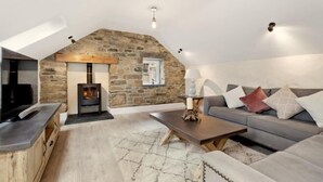An attic room with a stone wall, a fireplace, a sofa, a TV, and a coffee table. - Awel y Môr - 3 Bedroom Cottage - Whitesands (Saint Davids)
