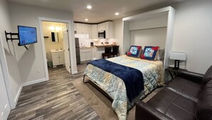 A compact bedroom with a bed, a brown leather sofa, a TV mounted on the wall, and a kitchenette in the background. - Back Beach Retreat - Car Rental Option (Sitka)