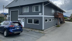 A two-story building with a grey exterior, numbered 1314, featuring a garage door, multiple windows, and a wooden staircase leading to an upper level. - Back Beach Retreat - Car Rental Option (Sitka)