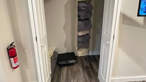 An open closet with hanging clothes and a folded blanket. A fire extinguisher is mounted on the wall. - Back Beach Retreat - Car Rental Option (Sitka)