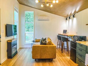 Un espacio habitable compacto con sofá, televisión, kitchenette y un ventanal amplio.