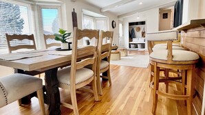 Een eetgedeelte met een houten tafel en stoelen, een hanglamp en uitzicht naar buiten door grote ramen.
