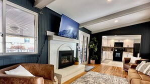 Een moderne woonkamer met een open haard, een televisie erboven en een keuken op de achtergrond.