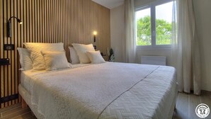 A neatly arranged bedroom with a large bed, a wooden slat wall, a window with curtains, and a nightstand with a lamp.