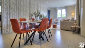 A modern dining area with a glass table and orange chairs, a sofa, and a decorative wall.