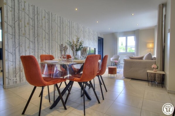 A modern dining area with a glass table and orange chairs, a sofa, and a decorative wall.