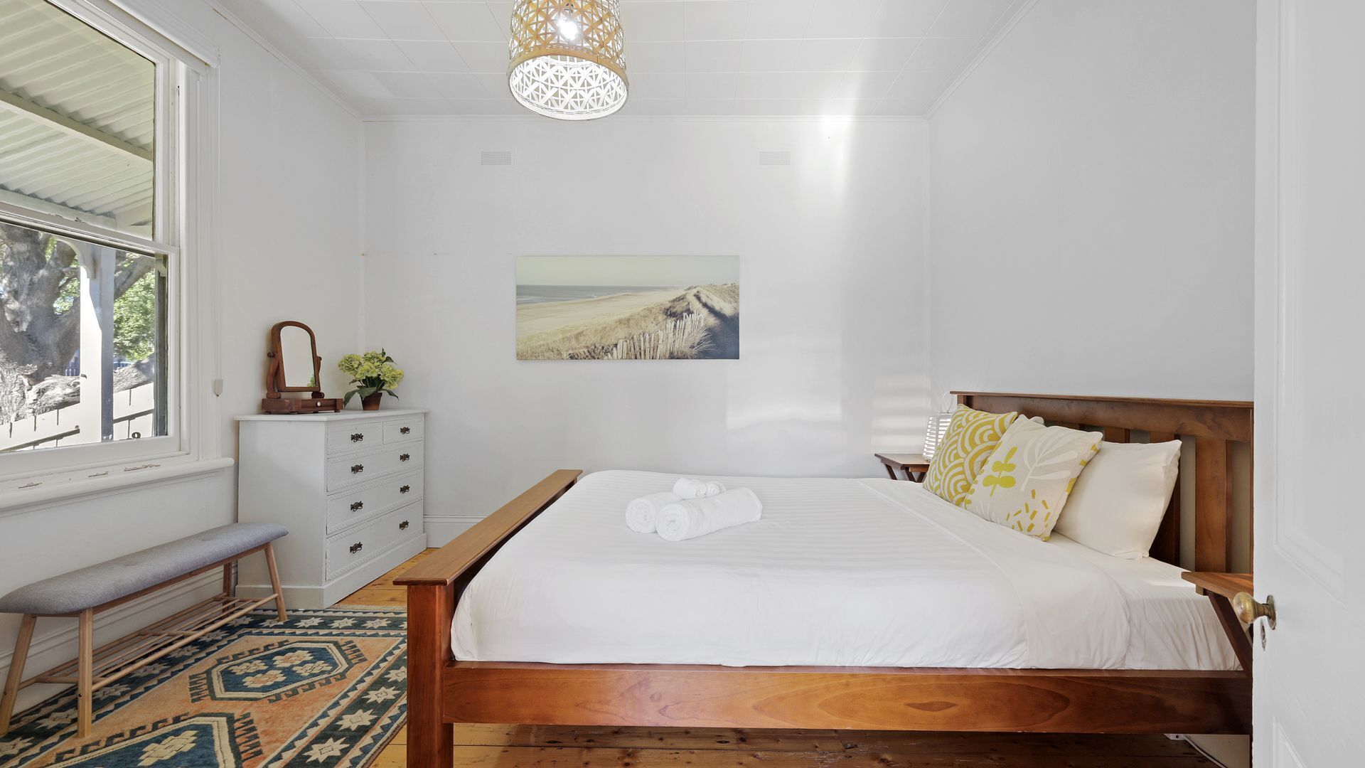 A bedroom with a wooden bed, white bedding, a bench, a dresser, and a wall-mounted picture.