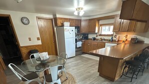A modern kitchen with wooden cabinets, a central island, and a dining area with a glass table and chairs. - Rustic Ridge (Gregory)