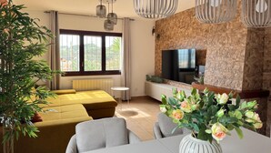 A modern living room with a sofa set, a dining table with chairs, a TV mounted on a stone wall, and a large window with a view.