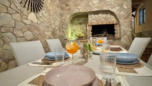 A stone-walled dining area with a fireplace, white chairs, and a table set for two with plates, glasses, and a fruit drink.