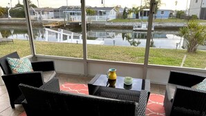 Un espacio de estar al aire libre con techo, mobiliario de mimbre y vista a un cuerpo de agua y casas al fondo.