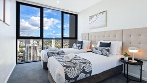 Une chambre d’hôtel moderne avec deux lits, une grande fenêtre donnant sur la ville, une tête de lit capitonnée et une table de chevet avec une lampe.