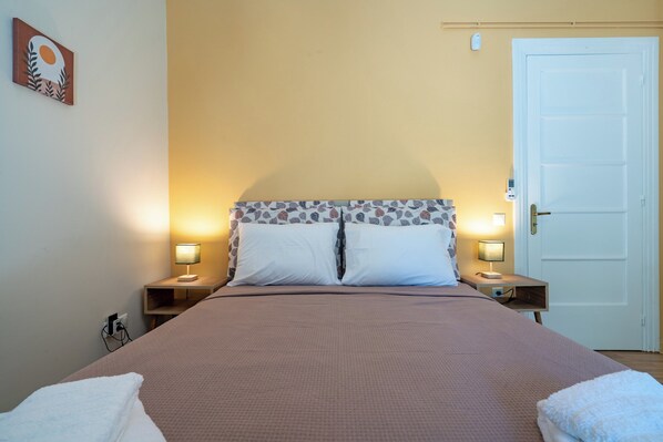 A neatly made bed with a brown bedspread and white pillows, flanked by two bedside tables with lamps, a white door, and a wall-mounted light fixture. - Monastiraki Aélia Apartment by ΜΡS (Athina)