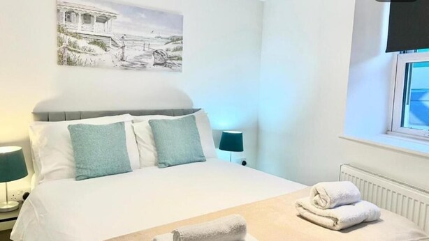 A neatly made bed with white and teal pillows, a bedside lamp, and a rolled towel on the bed. - Warmley Flat by Pulse Properties - close to Bristol Centre, Bath and Cotswolds. (Warmley)