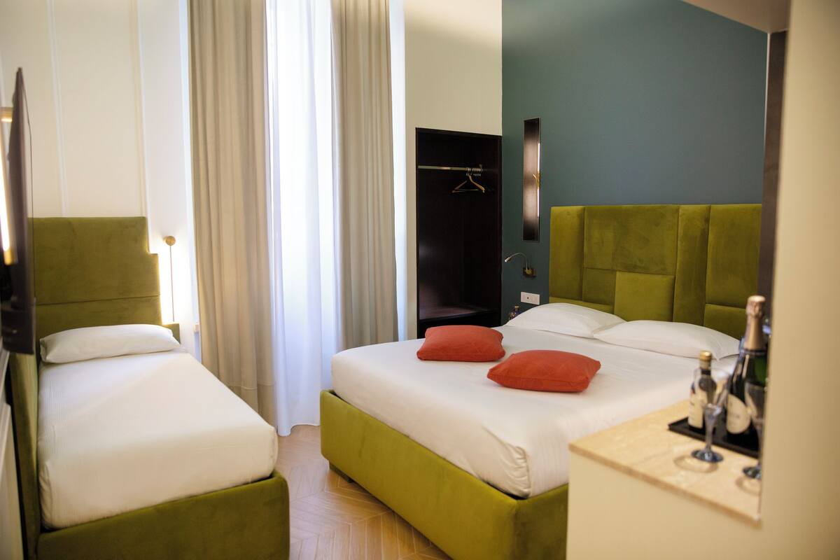 A hotel room with two single beds, a green headboard, a bedside table with a bottle and glass, and a hanging coat rack.