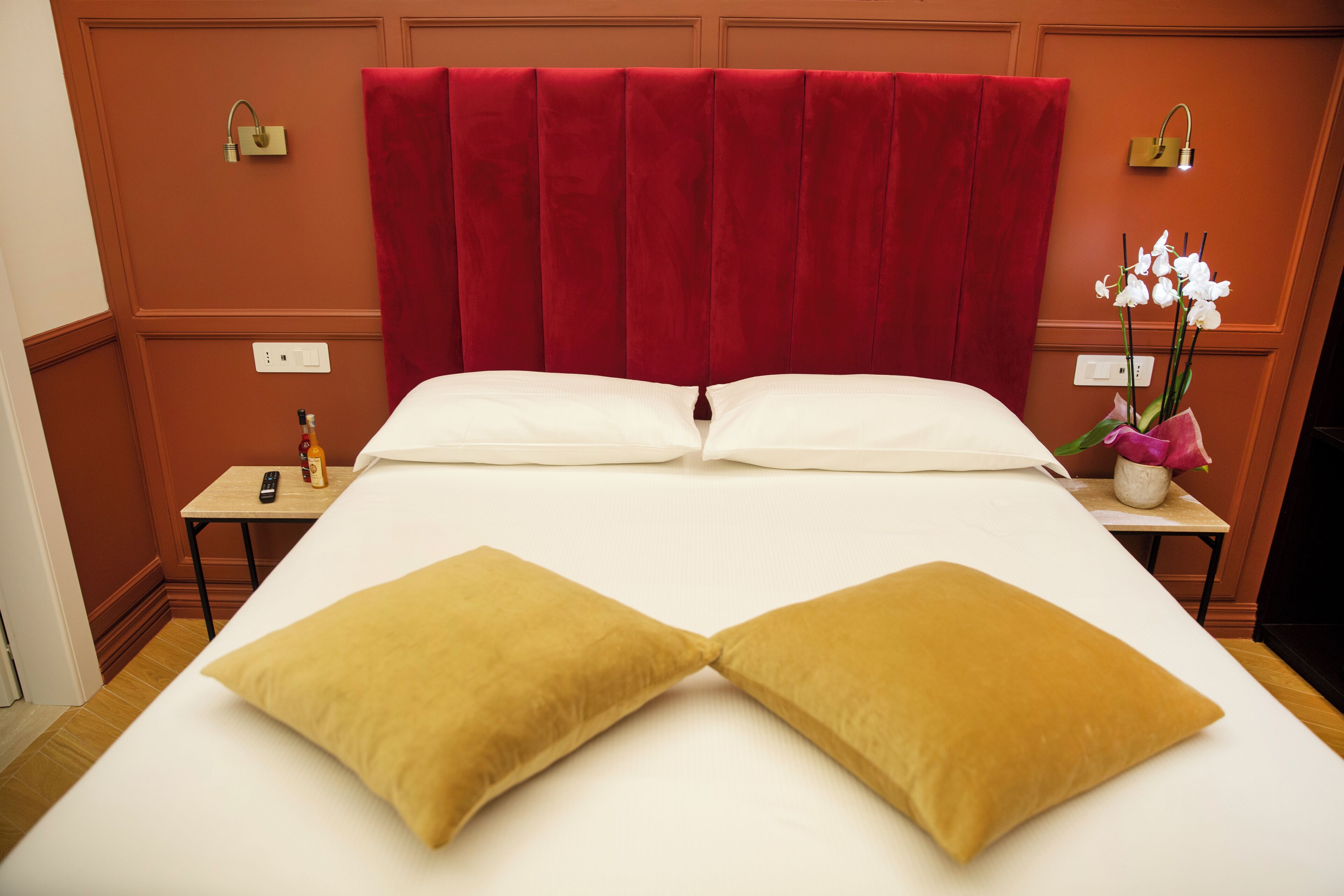 Quarto de hotel com uma cama grande, duas almofadas, mesa de cabeceira com uma garrafa e controle remoto, e um vaso com flores.