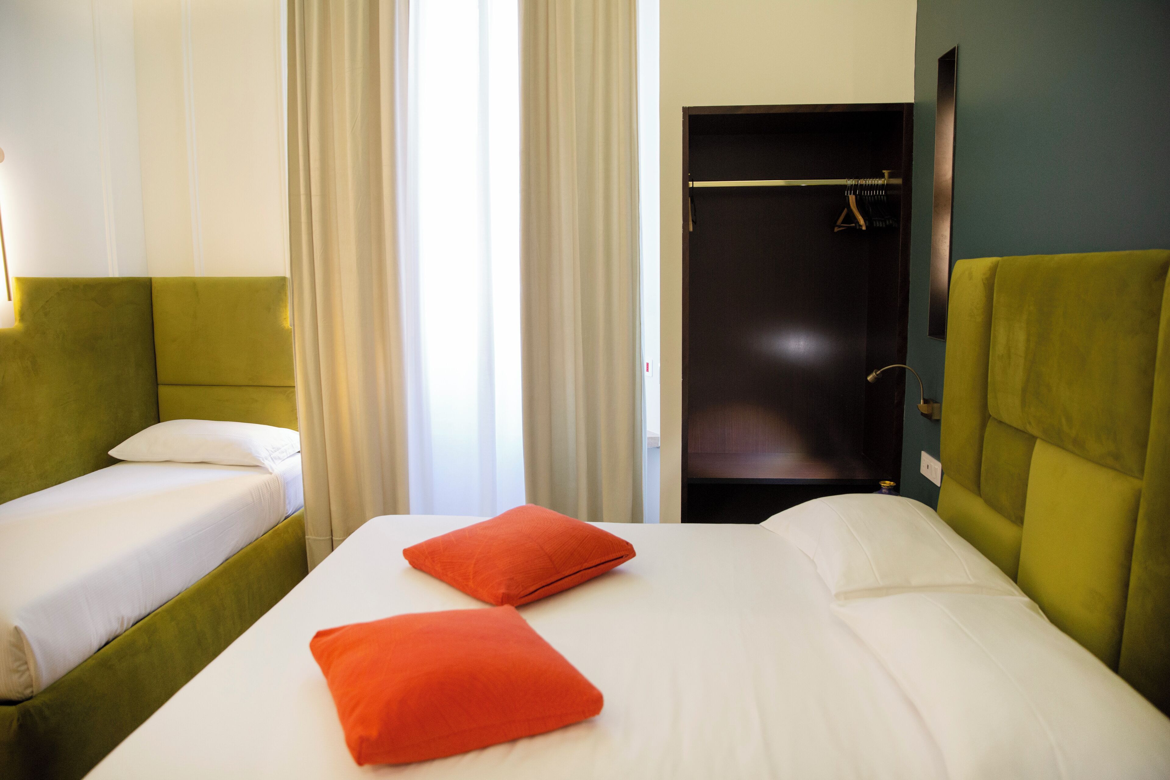 Une chambre d’hôtel avec un lit, deux oreillers, une tête de lit verte, une armoire et une fenêtre avec des rideaux.