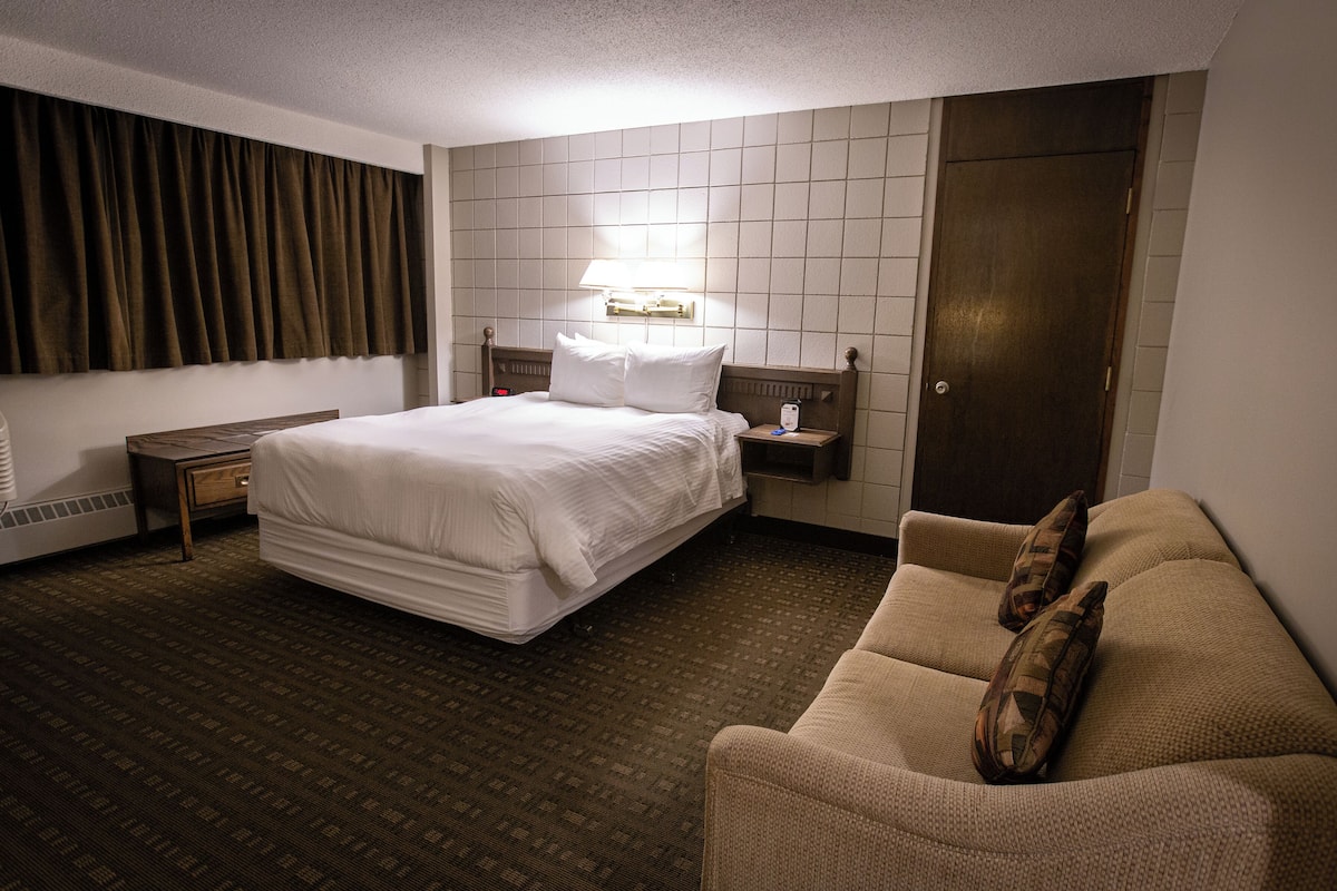 A hotel room with a bed, a nightstand, a lamp, a sofa, and a door.
