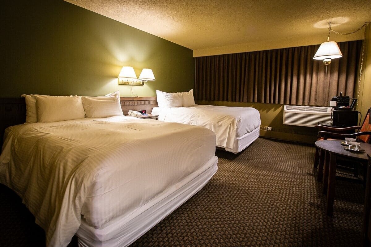 A hotel room with two beds, a desk, a chair, a lamp, and an air conditioning unit.