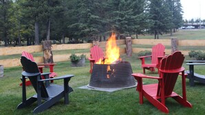 A fire pit with red and black Adirondack chairs around it, set in a grassy outdoor area with trees in the background. - Grande Cache Hotel (Grande Cache)