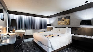 A modern hotel room with a large bed, a desk with a chair, a desk lamp, a wall-mounted TV, and a painting above the bed. - Grande Cache Hotel (Grande Cache)