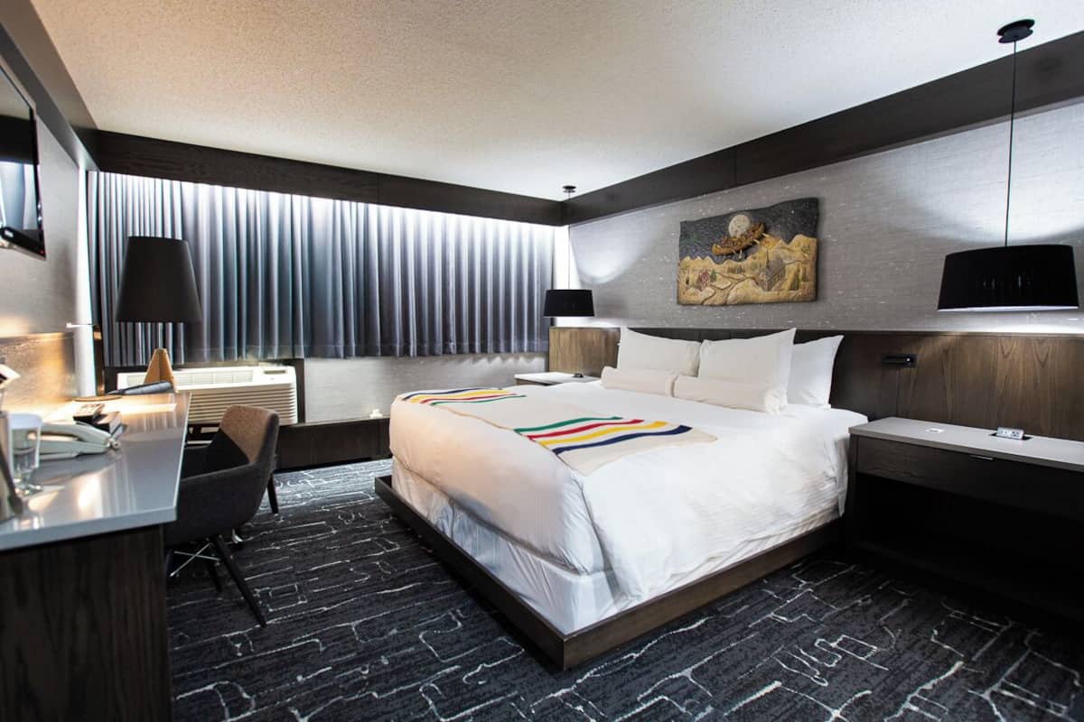 A modern hotel room with a large bed, a desk with a chair, a desk lamp, a wall-mounted TV, and a painting above the bed.