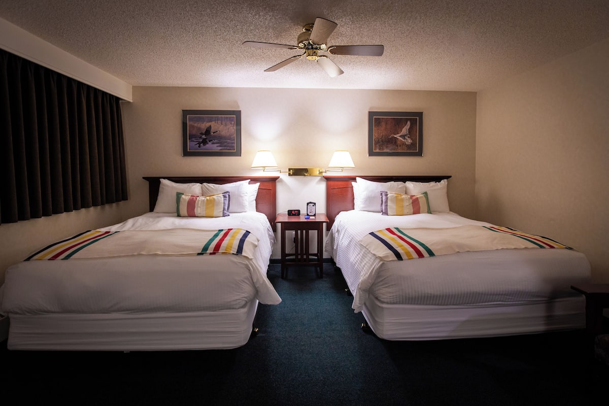 A hotel room with two beds, a ceiling fan, two wall-mounted lamps, and artwork on the walls.