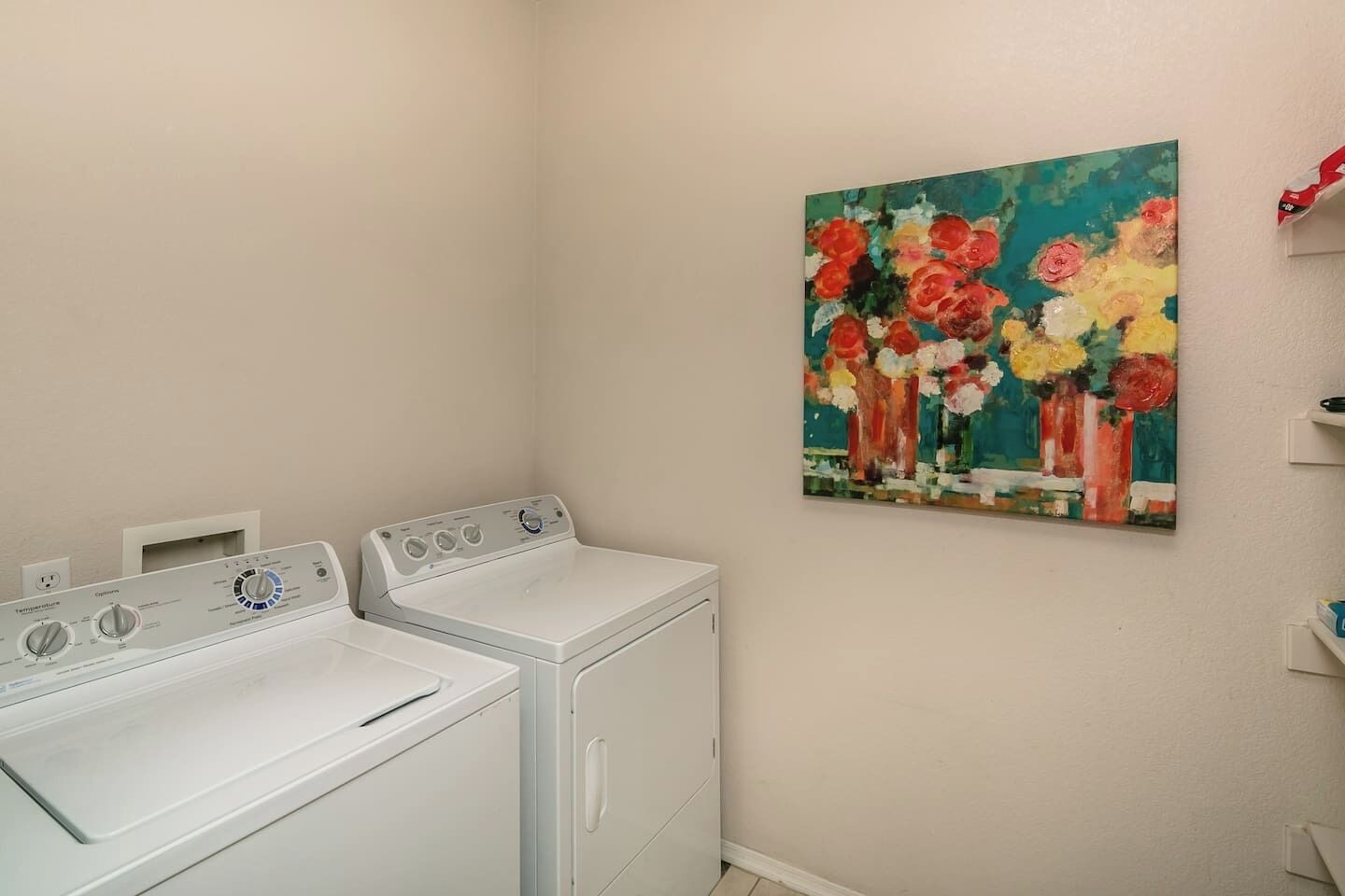 A laundry area with two white dryers and a painting of flowers on the wall.