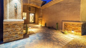 Une entrée bien éclairée, avec un chemin de pierre menant à une porte, le tout entouré de cactus et d’un mur de pierre.