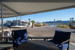 Uma área de estar coberta ao ar livre, com duas cadeiras e uma mesa, com vista para uma praia, palmeiras e montanhas ao longe.