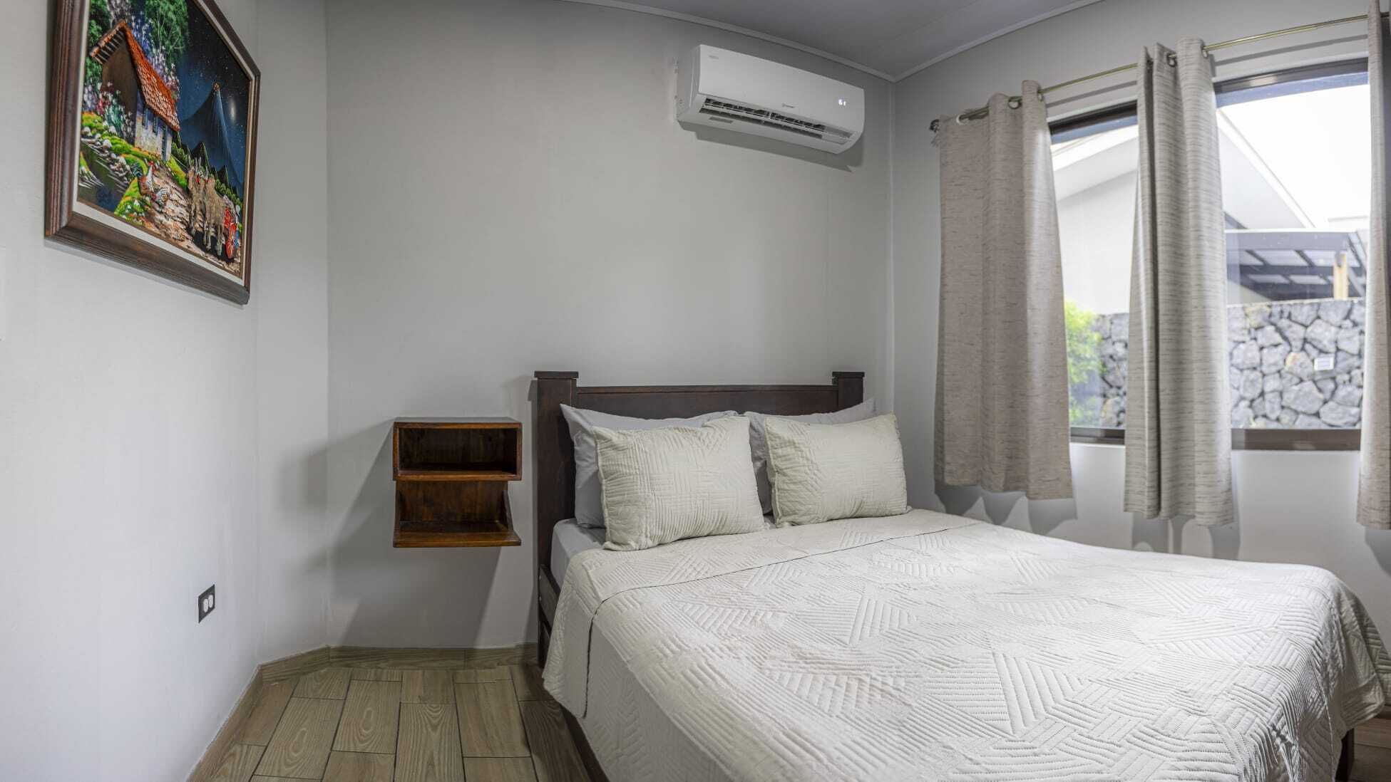 Une chambre à coucher comprenant un lit, une table de chevet, un tableau, un appareil de climatisation et une fenêtre avec des rideaux.