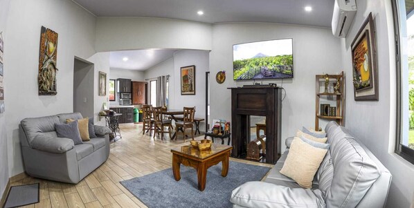 A spacious living room with a grey sofa, a wooden coffee table, and a fireplace. There is a dining area with wooden chairs and a table, and a kitchen in the background. - Casa Emunah (La Fortuna)